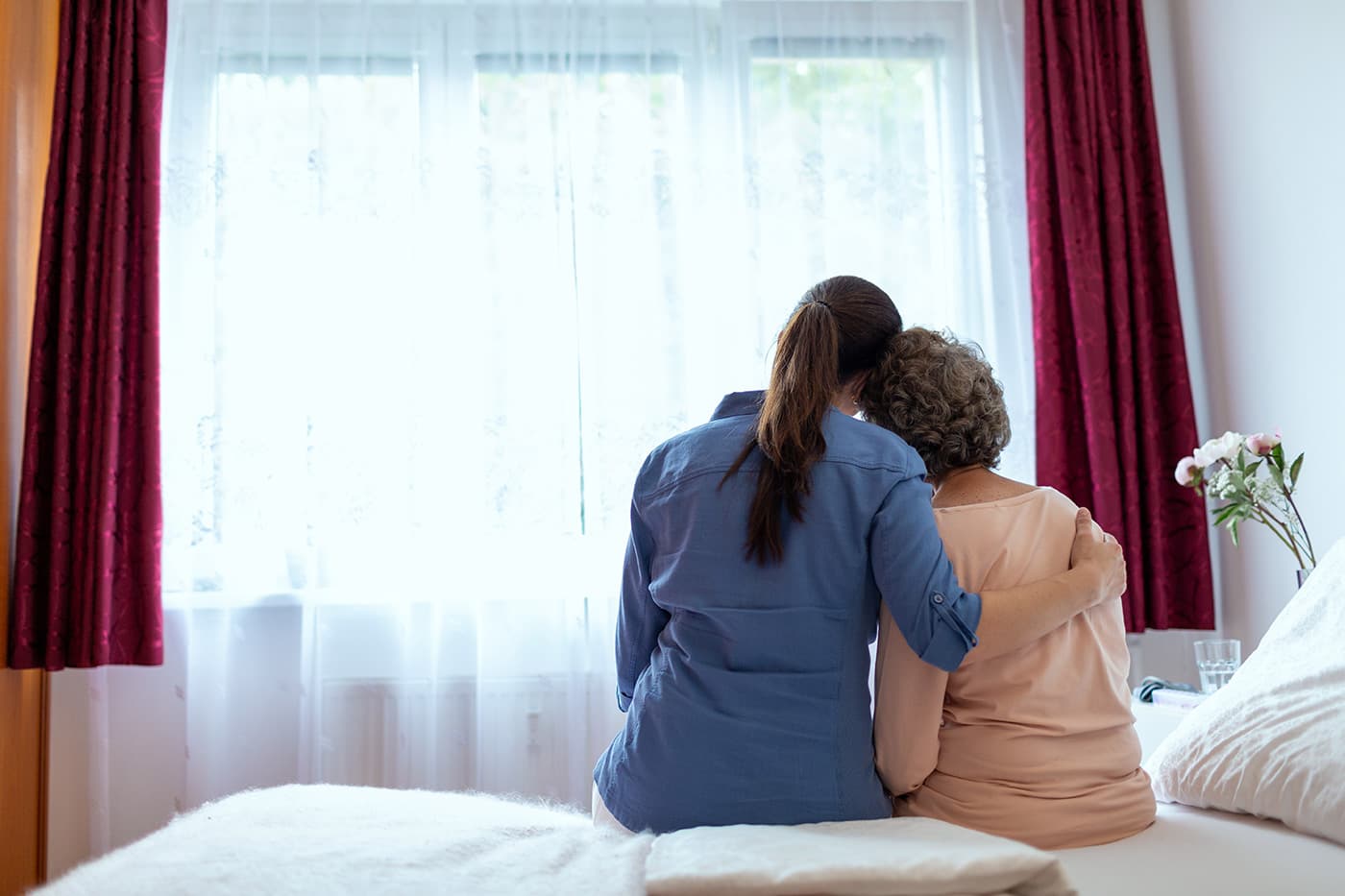 A younger person with their arm around an older adult, both seated on a bed and facing a window.