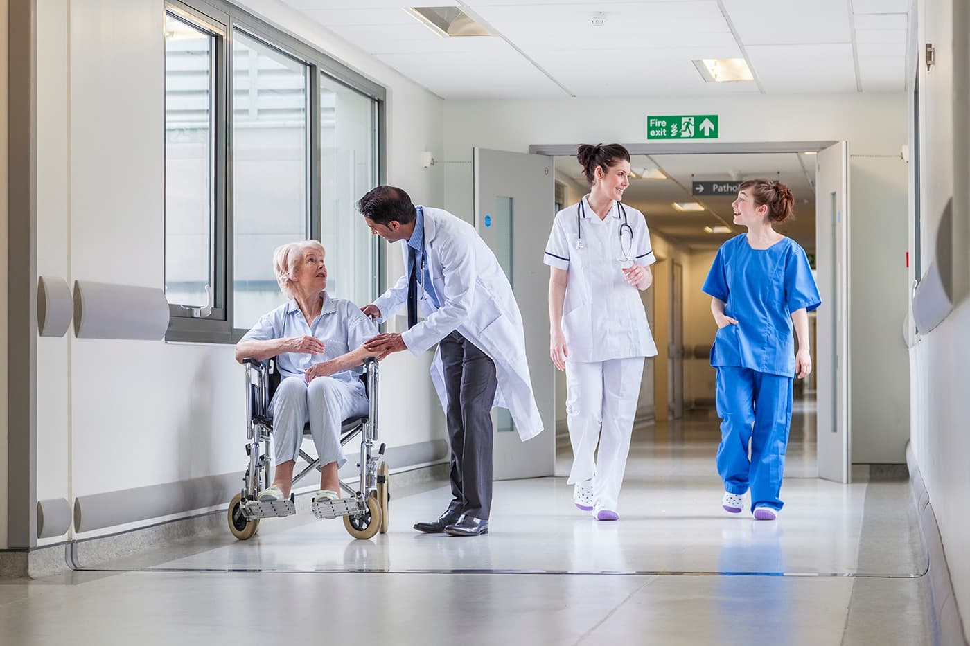 A doctor leans down to speak with an elderly patient seated in a wheelchair in a hospital hallway, while two nurses walk in the background.