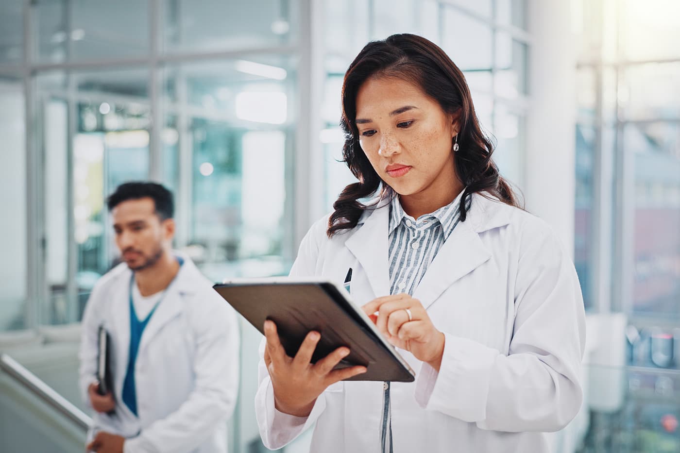 A female physician in a white coat reviews information on a tablet while standing in a bright hospital corridor.