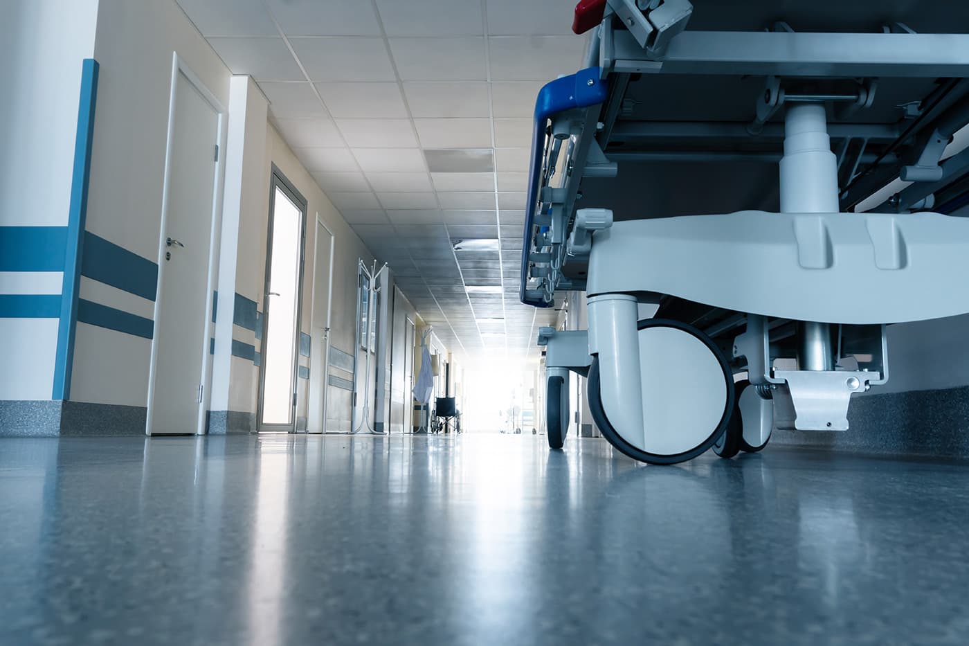 A low-angle view of a long hospital corridor, with a gurney in the foreground and a wheelchair visible in the distance near a bright window.