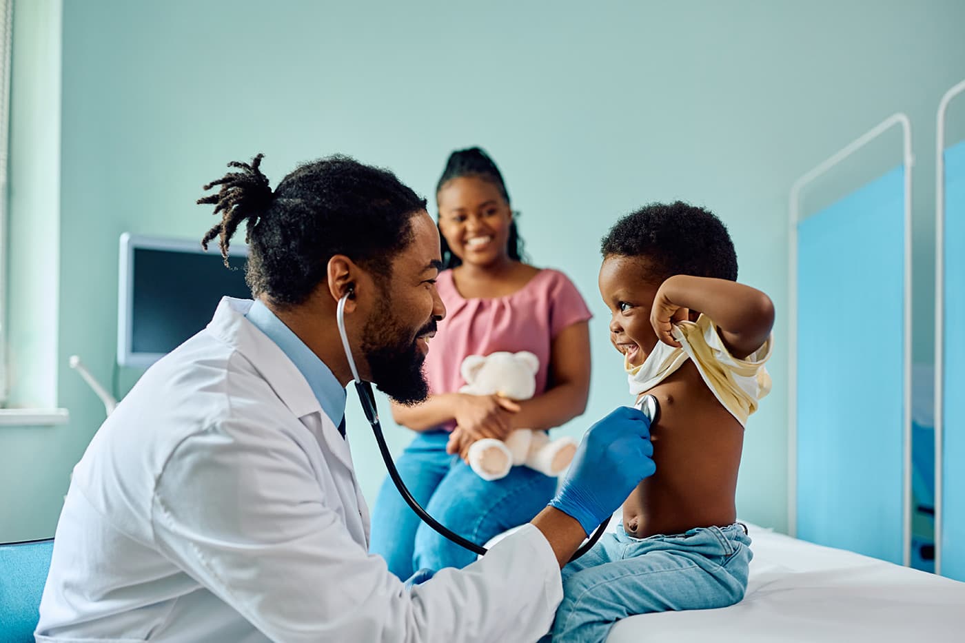 A smiling pediatrician uses a stethoscope to examine a young child, while the child's parent looks on in the background.