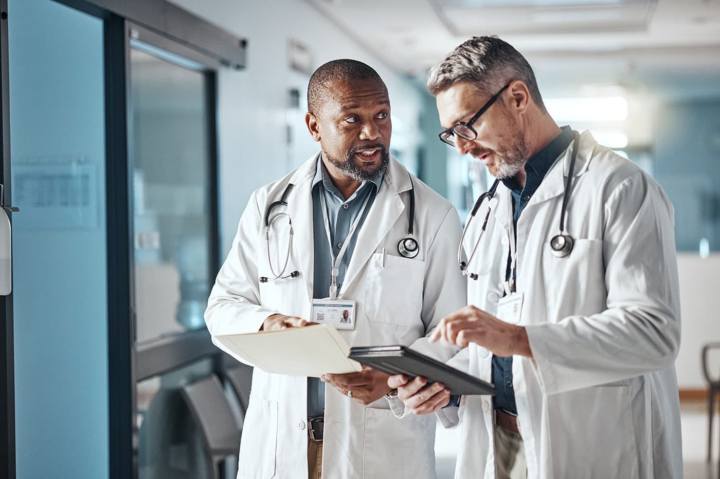 Two doctors in white coats standing in a hospital hallway, reviewing documents and a tablet together